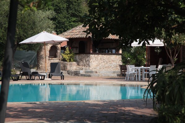 A swimming pool with lounge chairs and umbrellas, a stone structure with a tiled roof, and a table with chairs.