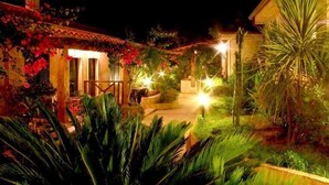 Un patio bien iluminado con un edificio que tiene un balcón y una zona cubierta, rodeado de abundante vegetación y plantas.