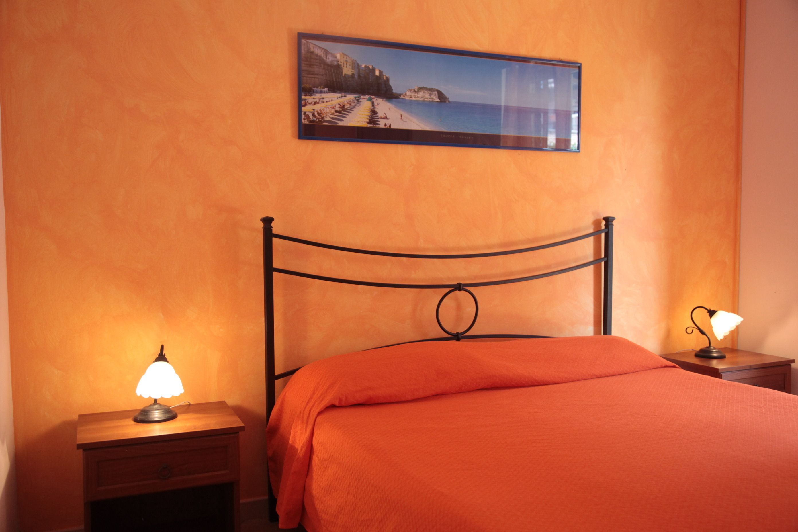 Un dormitorio con cama, mesita de noche con lámpara, un cuadro enmarcado en la pared y cabecera de madera.
