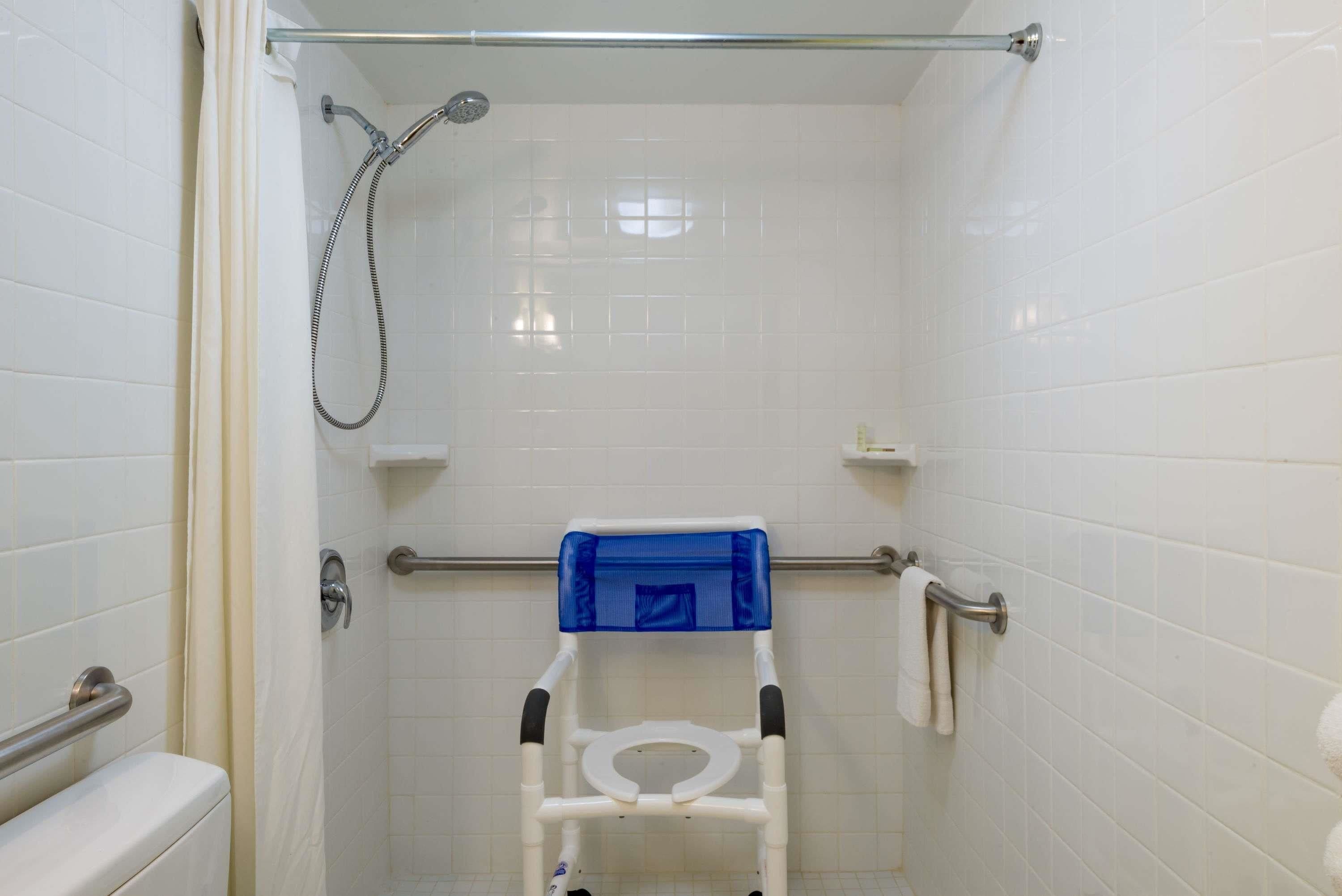A small, enclosed shower stall with a white tiled wall, a showerhead, a grab bar, and a blue safety seat.