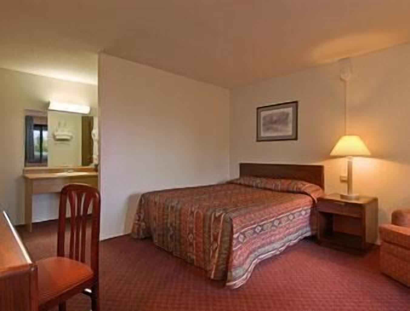 A hotel room with a bed, a desk, a chair, a nightstand, and a lamp.