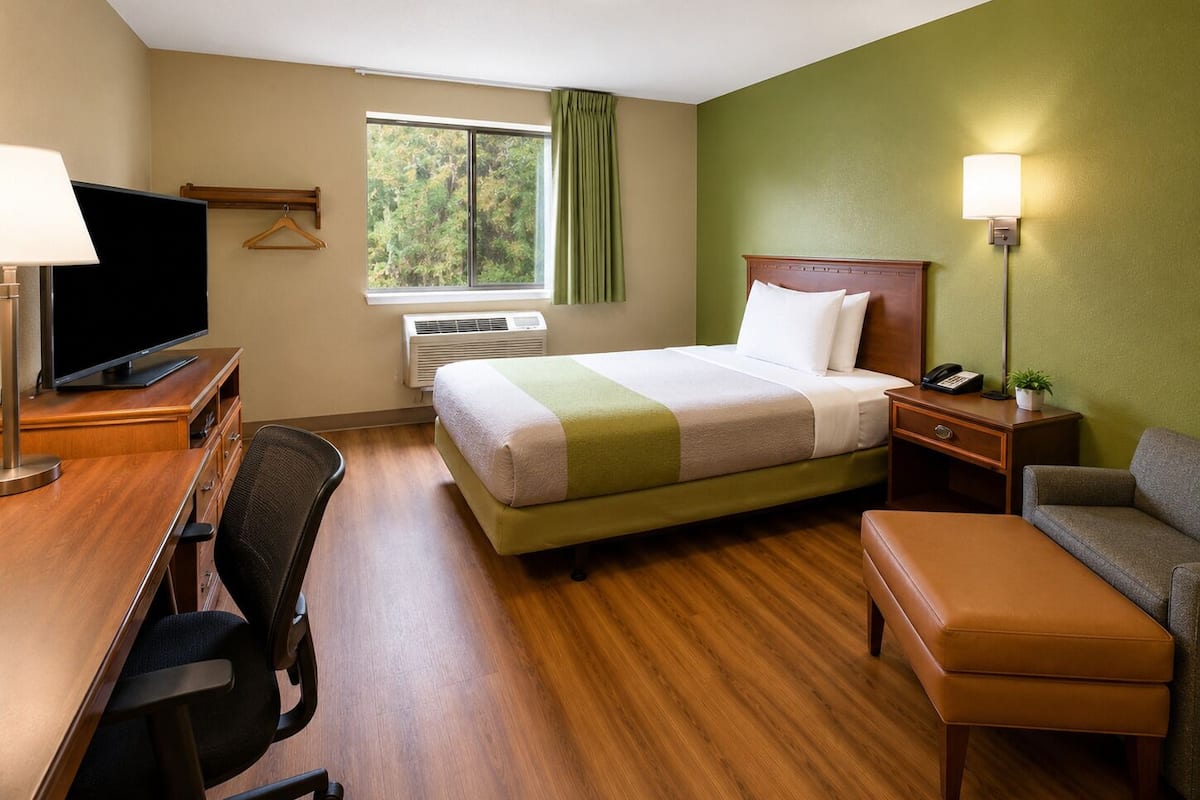 Habitación de hotel con cama, escritorio, silla, televisor y ventana con cortinas.