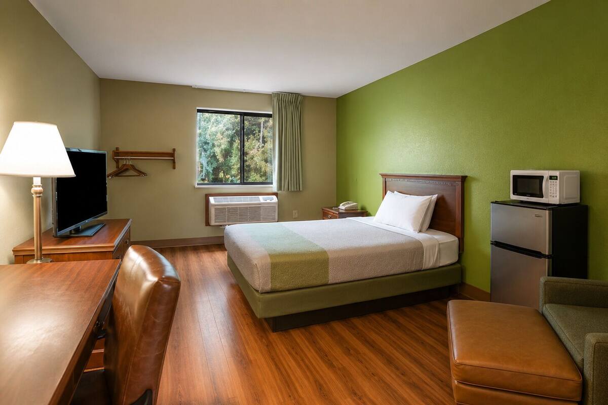 A hotel room with a bed, a desk with a lamp, a television, a refrigerator, and a window with curtains.