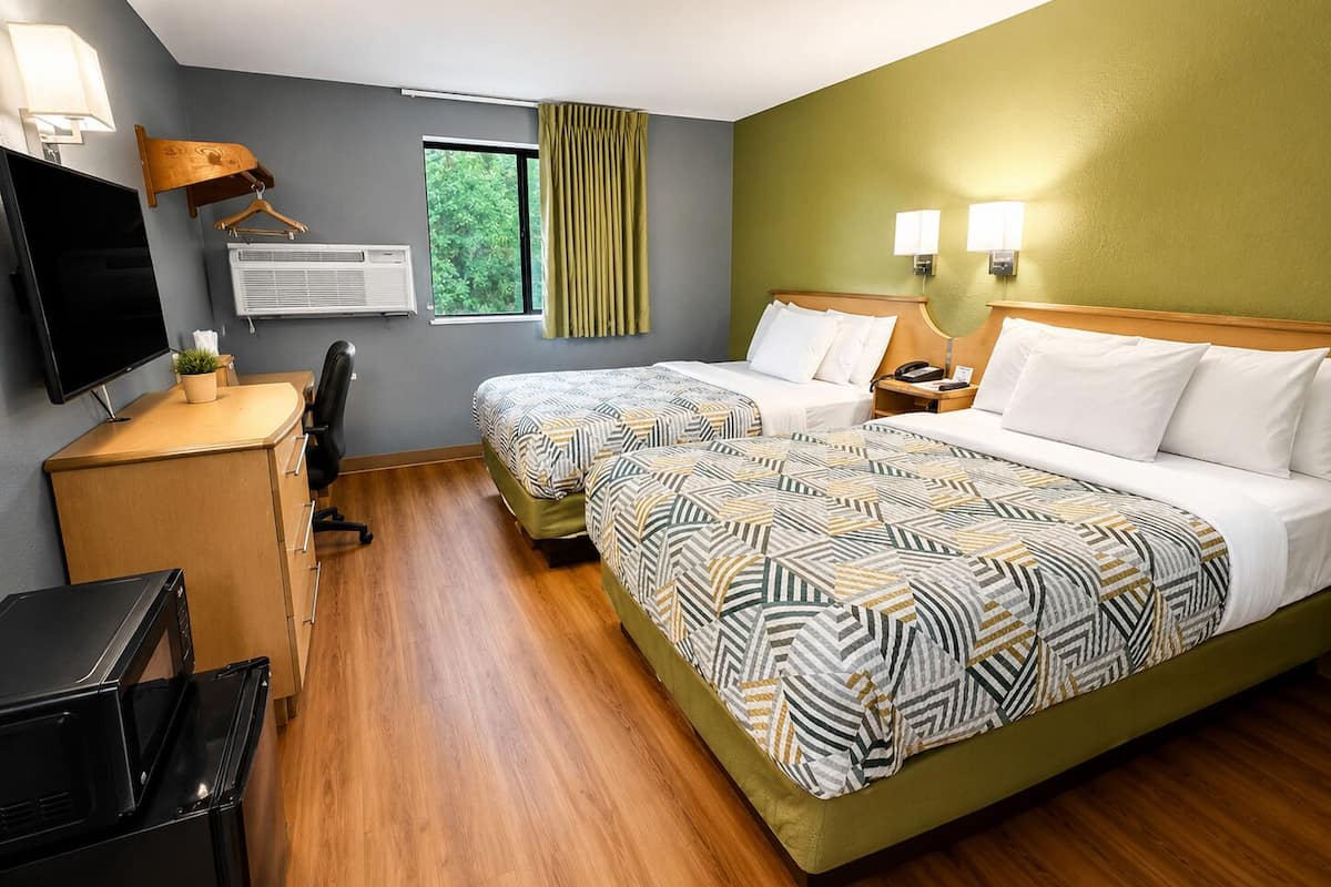 Habitación de hotel con dos camas, un escritorio con televisión, una silla, unidad de aire acondicionado y una ventana con cortinas.