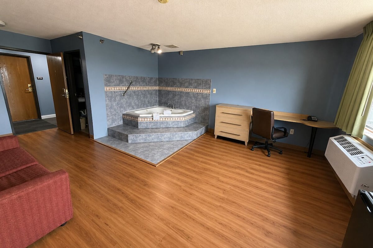 Una habitación con piso de madera, un sofá rojo, un escritorio, una silla, una bañera y una ventana con cortinas.