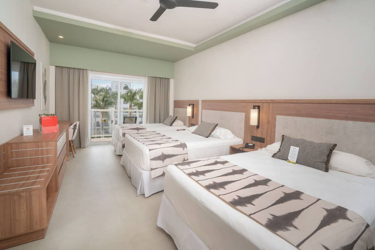A hotel room with two beds, a desk, a TV, and a balcony with a view of palm trees.