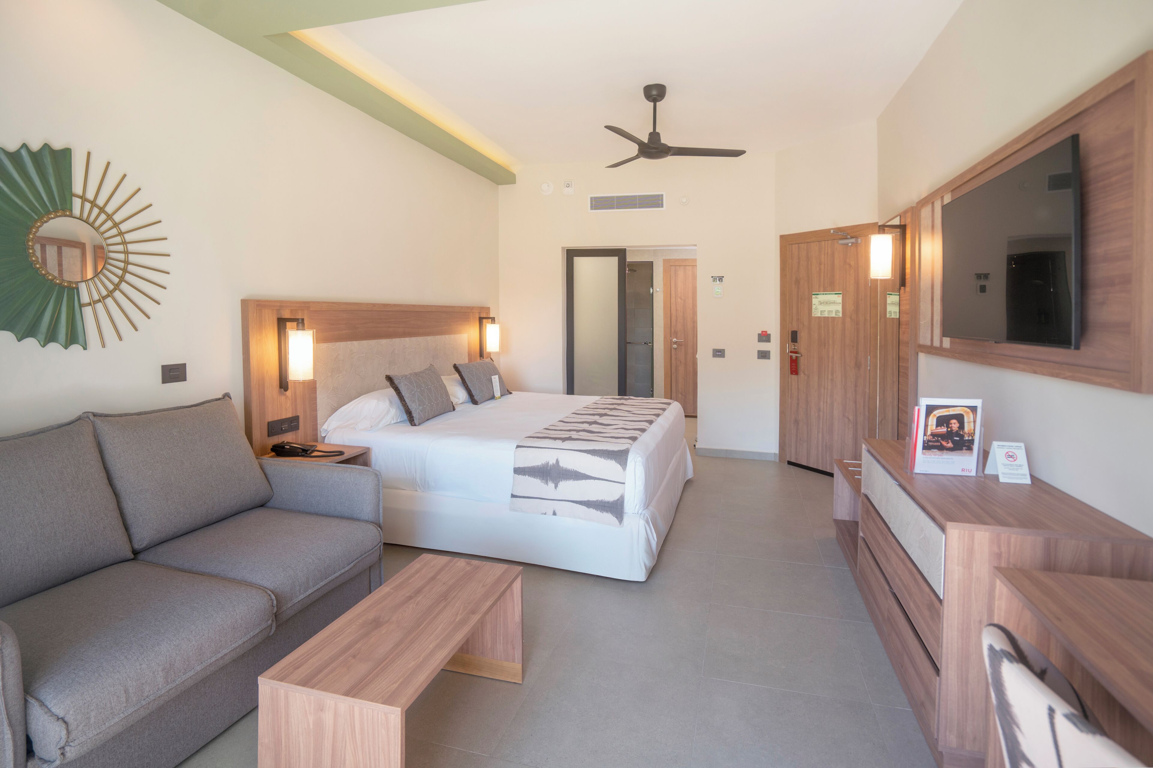 A hotel room with a bed, a sofa, a desk, a mirror, and a ceiling fan.