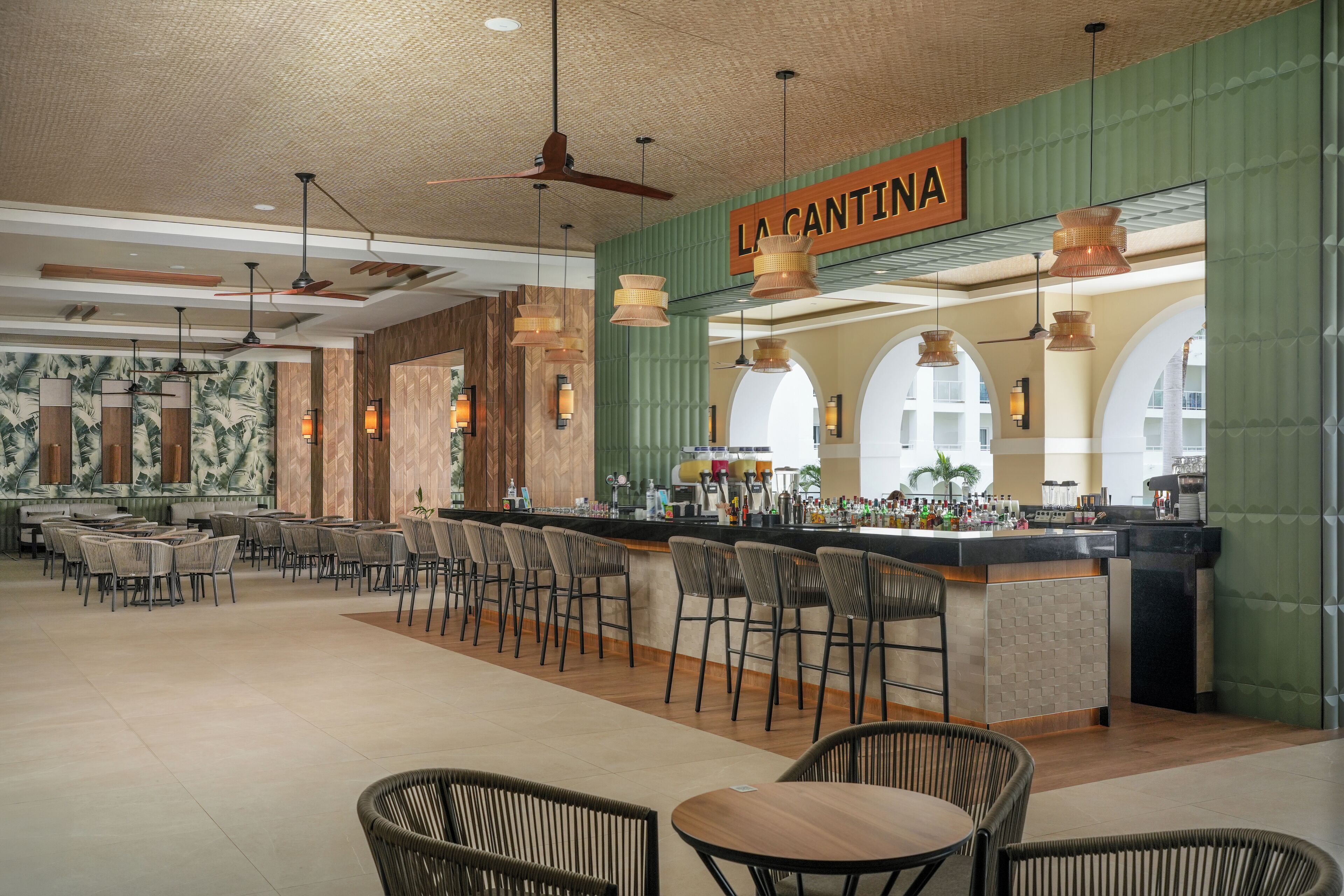 En rummelig bar med disk og siddepladser, et skilt der viser "LA CANTINA", og et glimt af køkkenet i baggrunden.