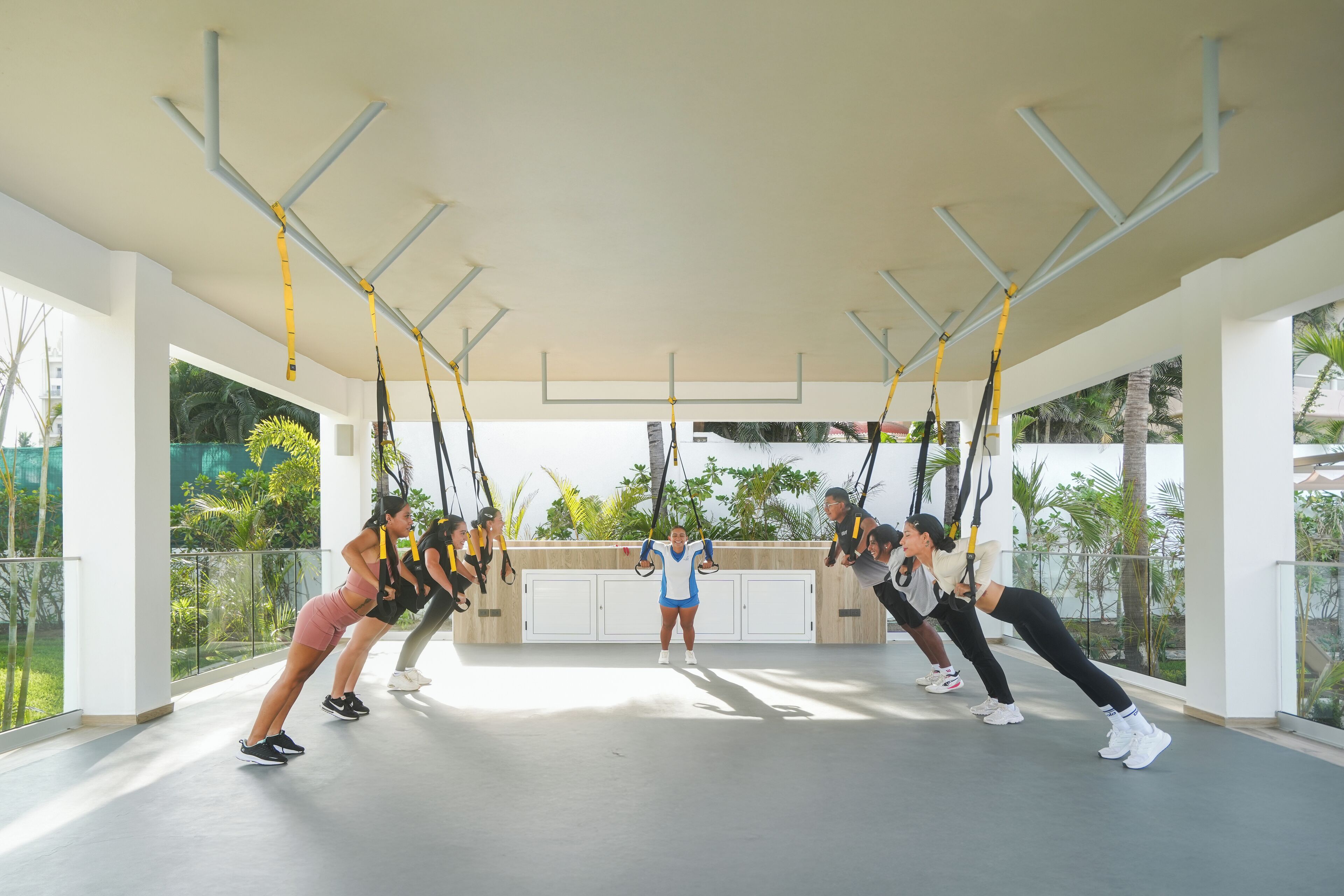 A group of people exercising with TRX suspension straps in a spacious, well-lit room with large windows and a view of greenery outside.