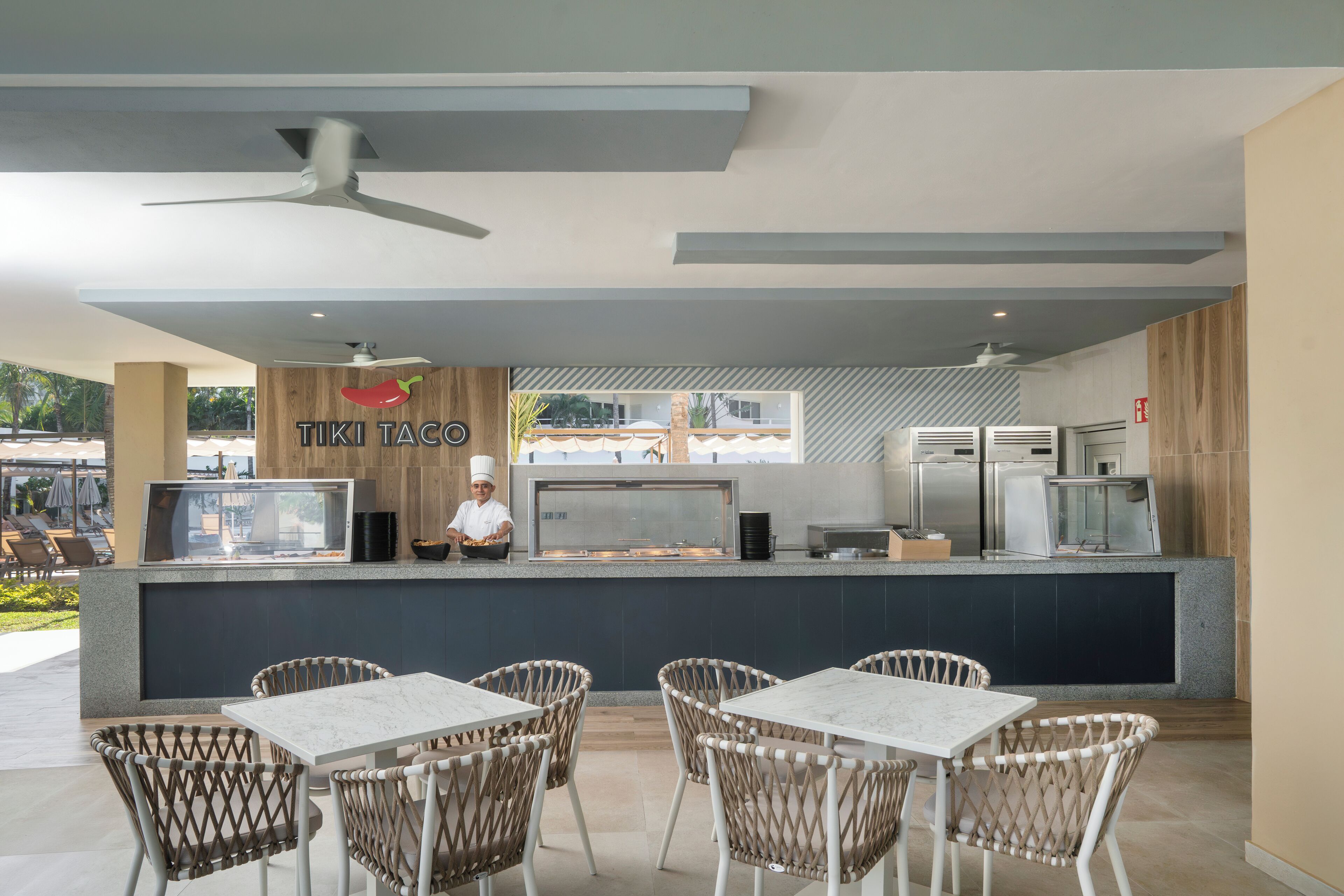 Un área de comedor al aire libre con sillas de mimbre y una mesa blanca, una barra con un chef, y vistas al exterior.
