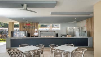 Un área de comedor al aire libre con sillas de mimbre y una mesa blanca, una barra con un chef, y vistas al exterior.