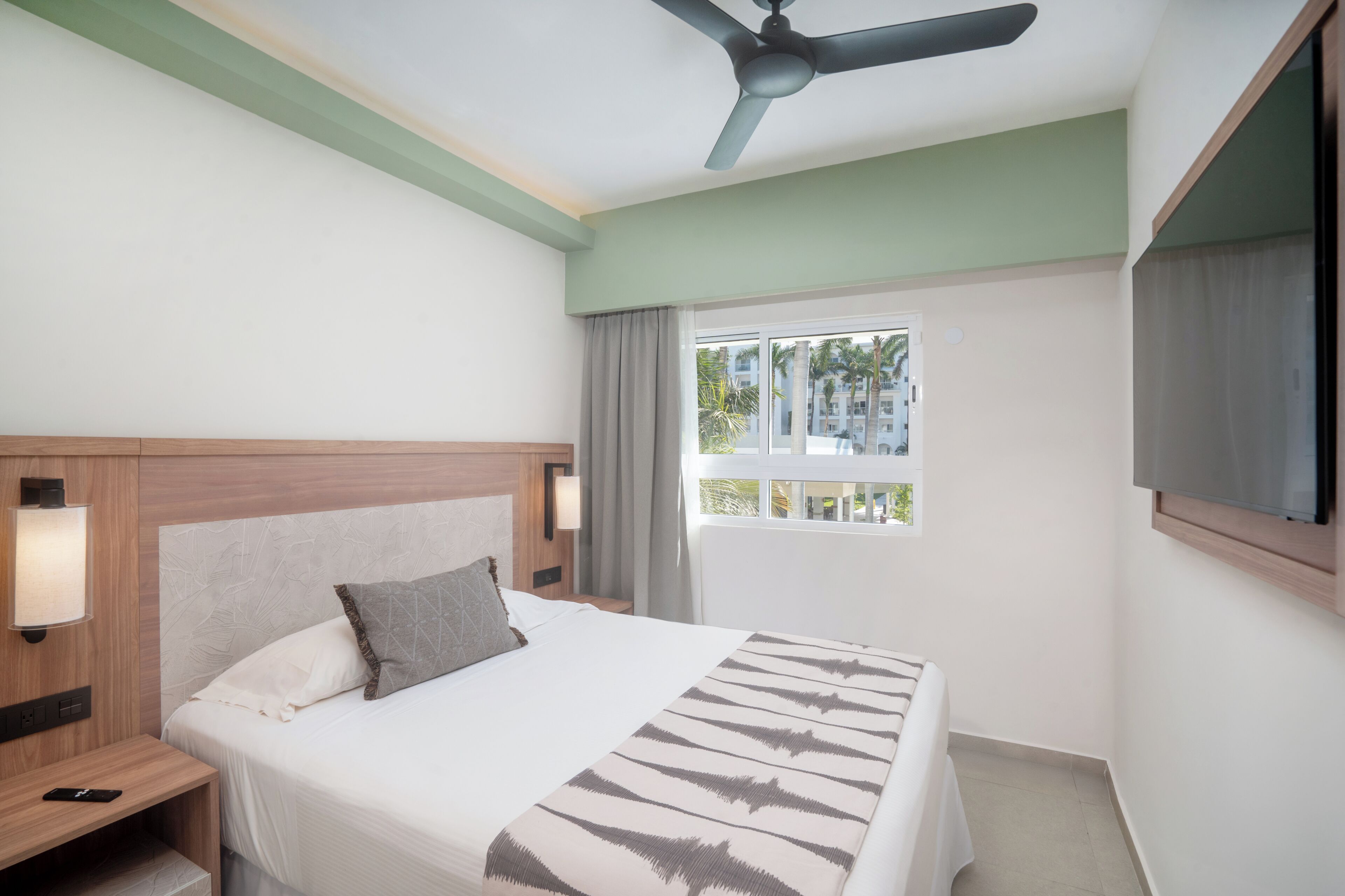 A hotel room with a bed, nightstand, ceiling fan, window with curtains, and a TV.
