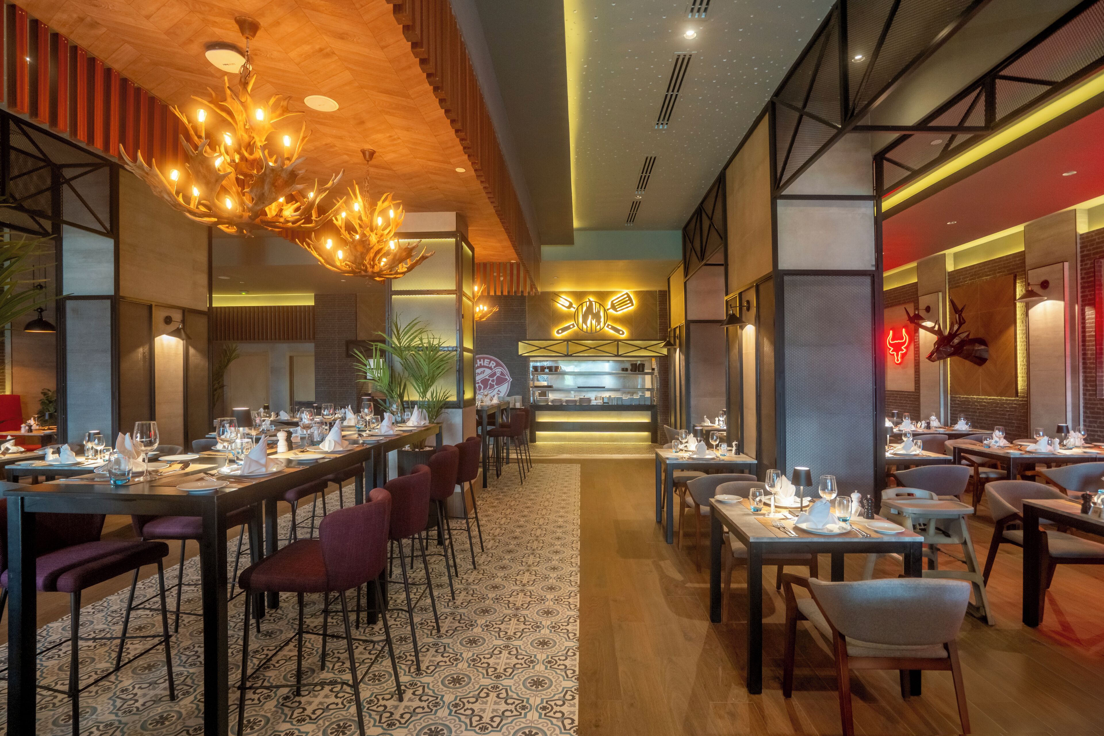 A modern restaurant with long communal tables, red upholstered chairs, and a patterned carpet.