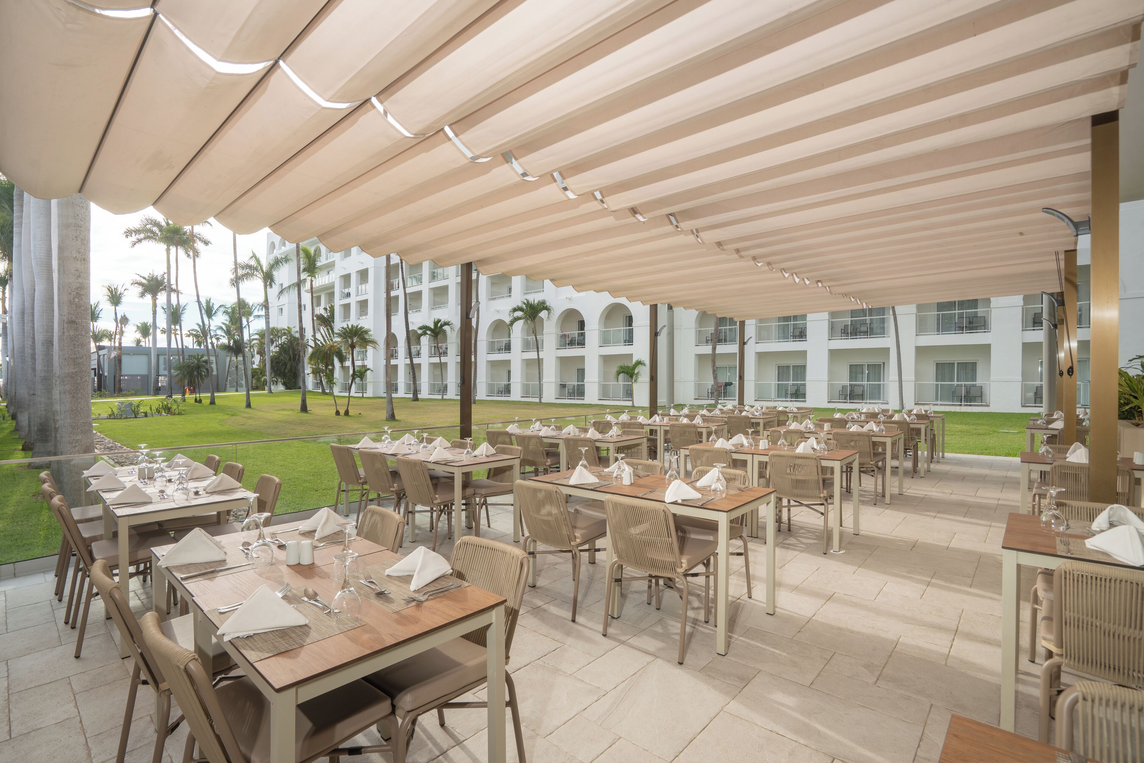 Un área de comedor al aire libre con mesas y sillas, una zona cubierta con techo y una vista de un edificio con varios balcones y palmeras al fondo.