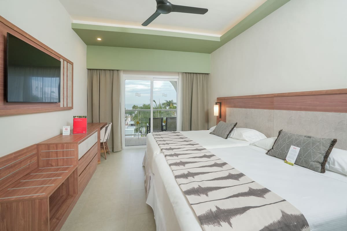A hotel room with a bed, a desk, a television, a ceiling fan, and a balcony with a view.