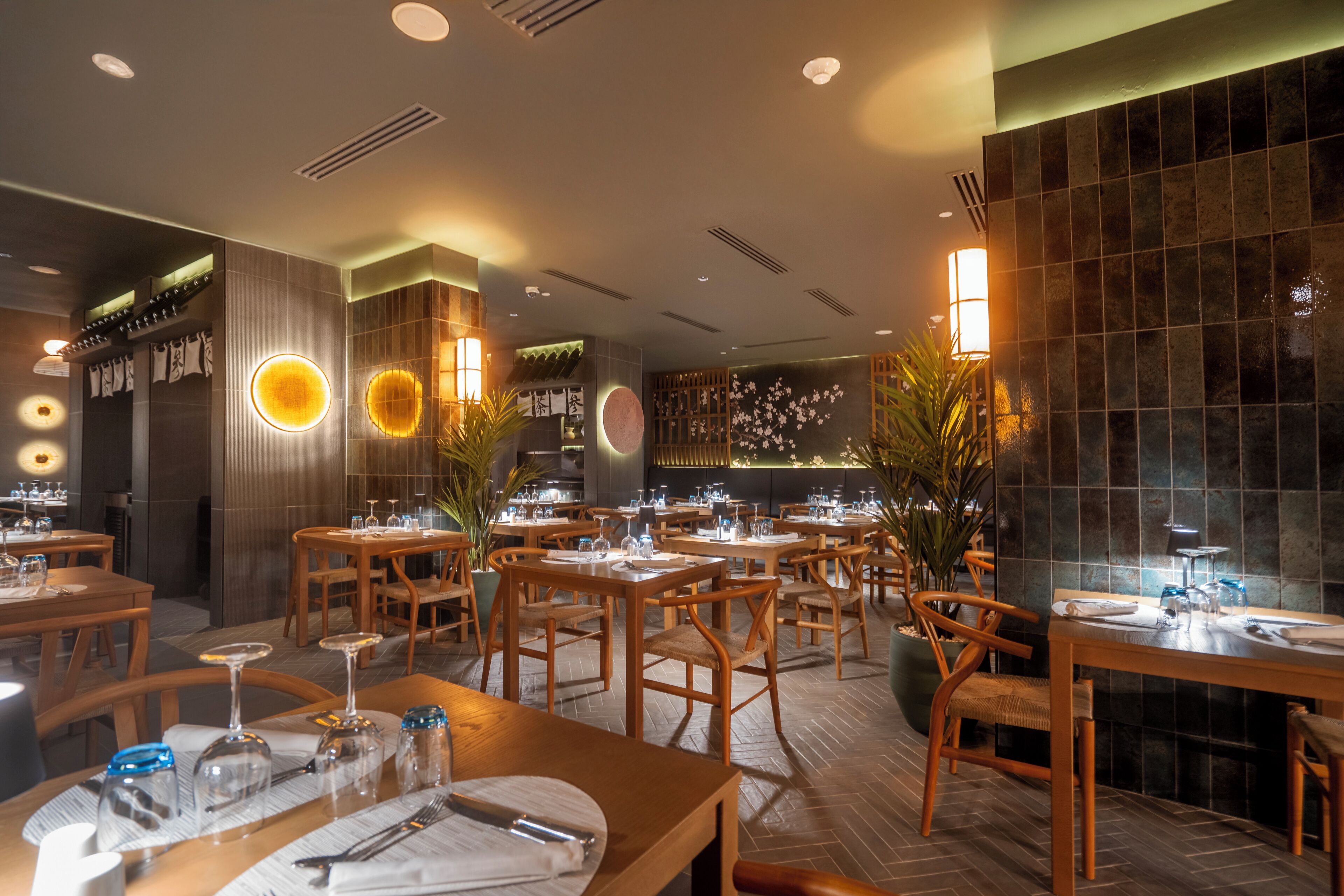 Un restaurante con mesas y sillas de madera, mesas redondas con centros de mesa, y una pared de azulejos con un motivo floral.