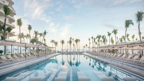 A swimming pool with lounge chairs and umbrellas, surrounded by palm trees and a clear sky. - Riu Flamingos - All Inclusive (Nuevo Nayarit)