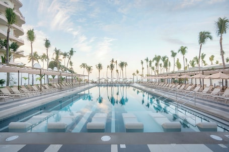 A swimming pool with lounge chairs and umbrellas, surrounded by palm trees and a clear sky.