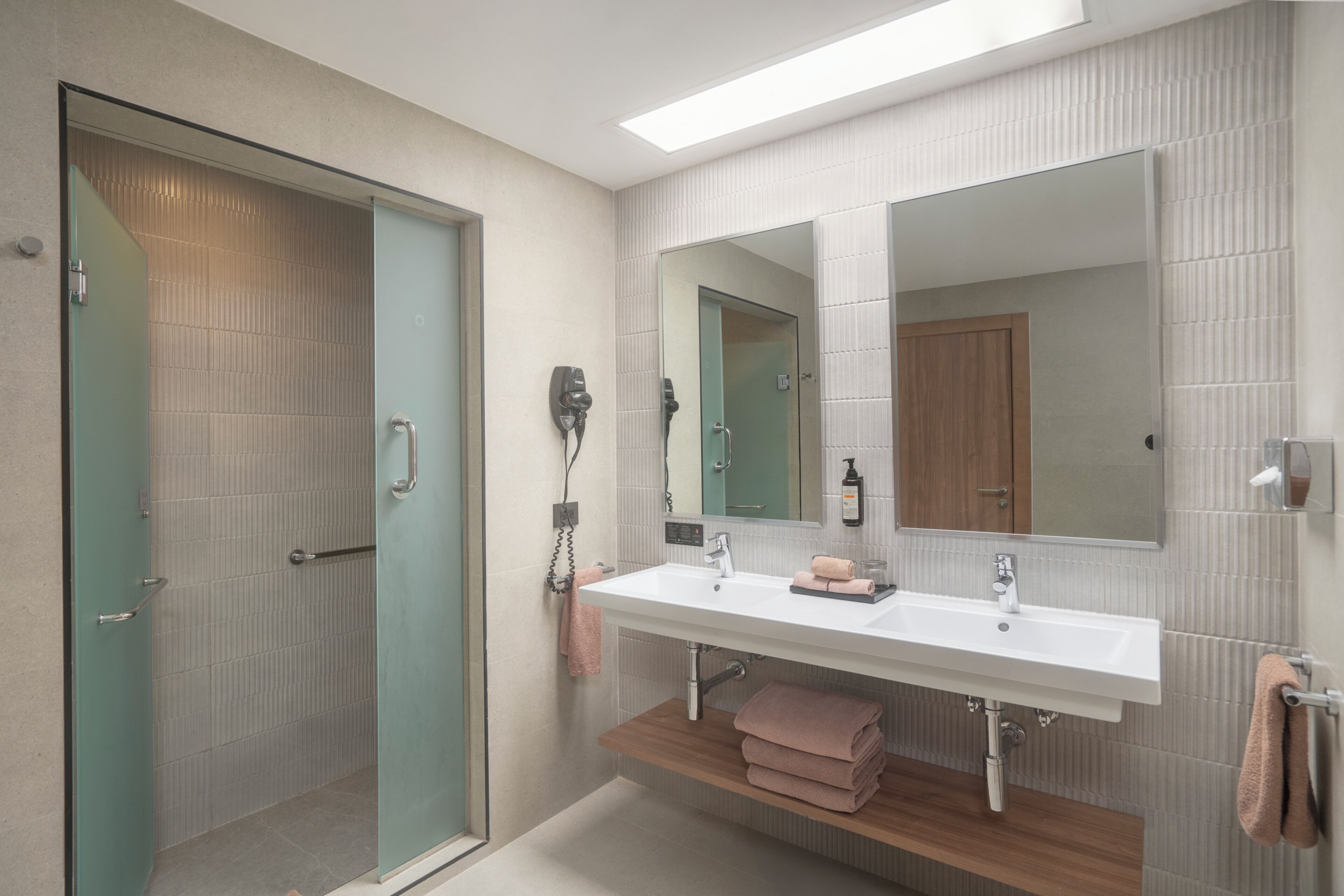 A modern bathroom with a glass shower, a white sink, a large mirror, and a wooden shelf.