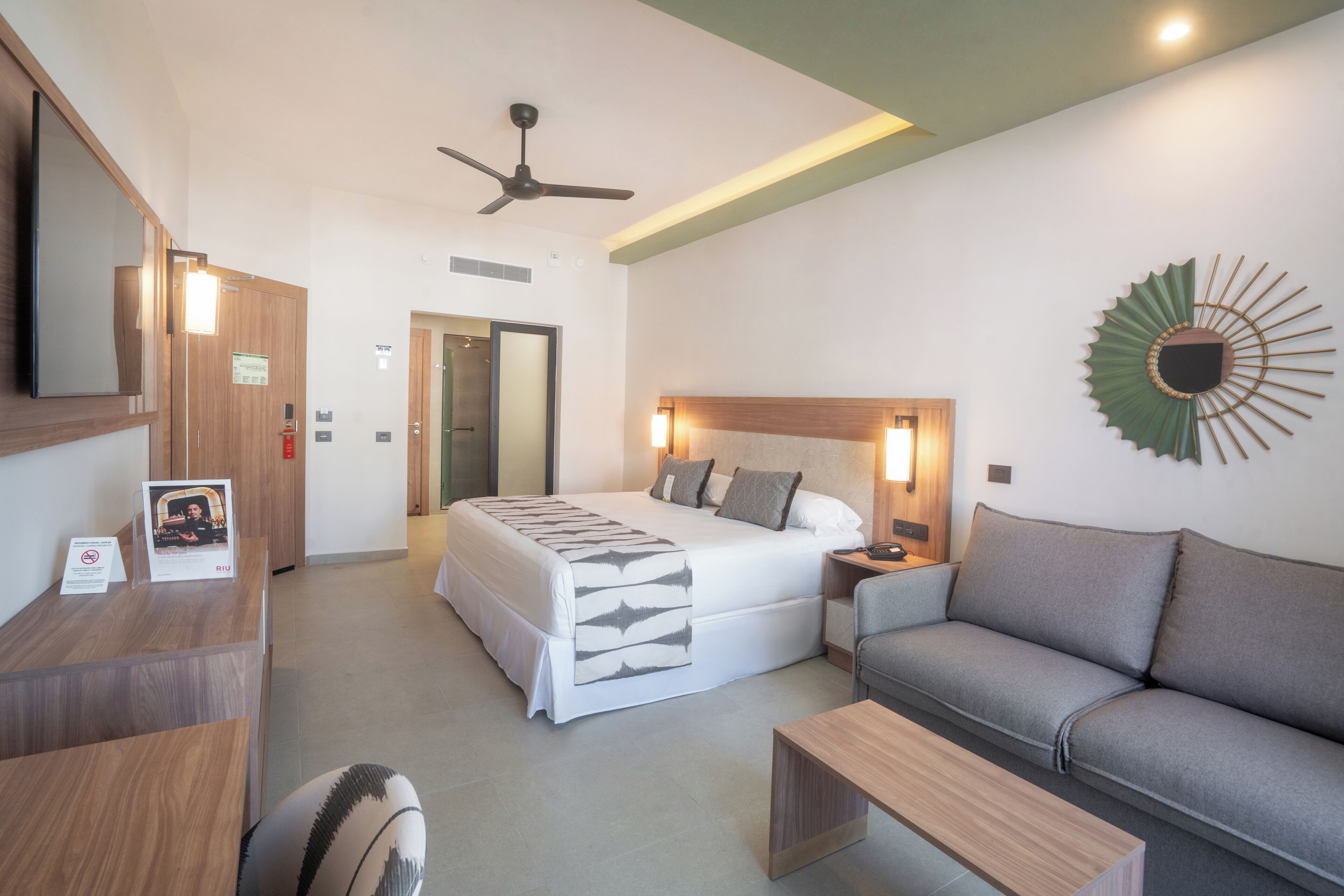 A hotel room with a bed, a sofa, a desk, a TV, a ceiling fan, and a decorative wall piece.