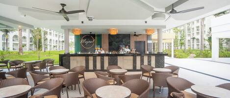 A bar area with wicker chairs and tables, a counter with a bartender, and a view of a building with columns and greenery.