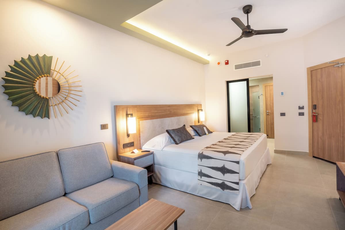 A hotel room with a bed, a sofa, a nightstand, a ceiling fan, and a wall-mounted decorative piece.