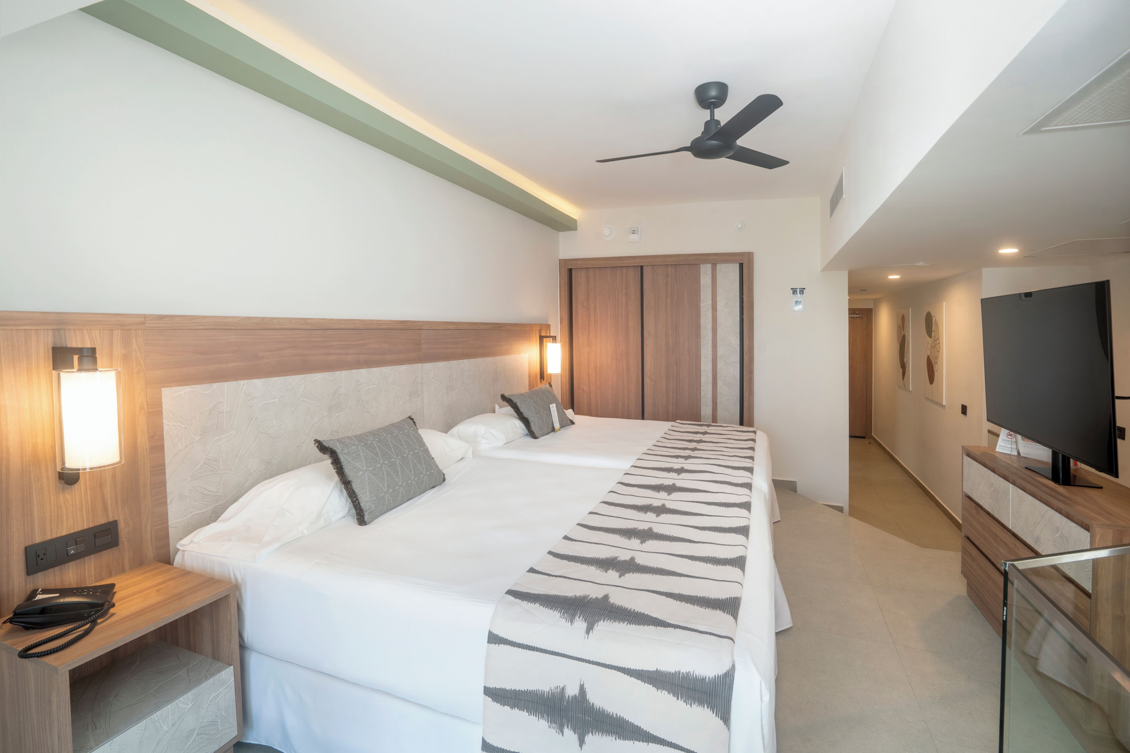 Una habitación de hotel con una cama grande, mesitas de noche, un ventilador de techo, televisor y armario.