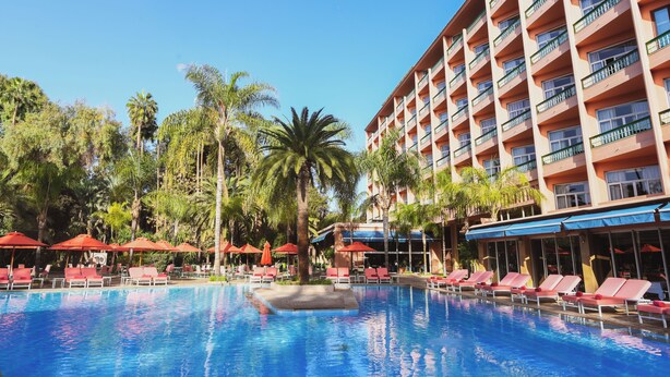 A hotel pool area with sun loungers and umbrellas, surrounded by palm trees and a multi-story building. - Es Saadi Marrakech Resort Hotel (Marrakech)
