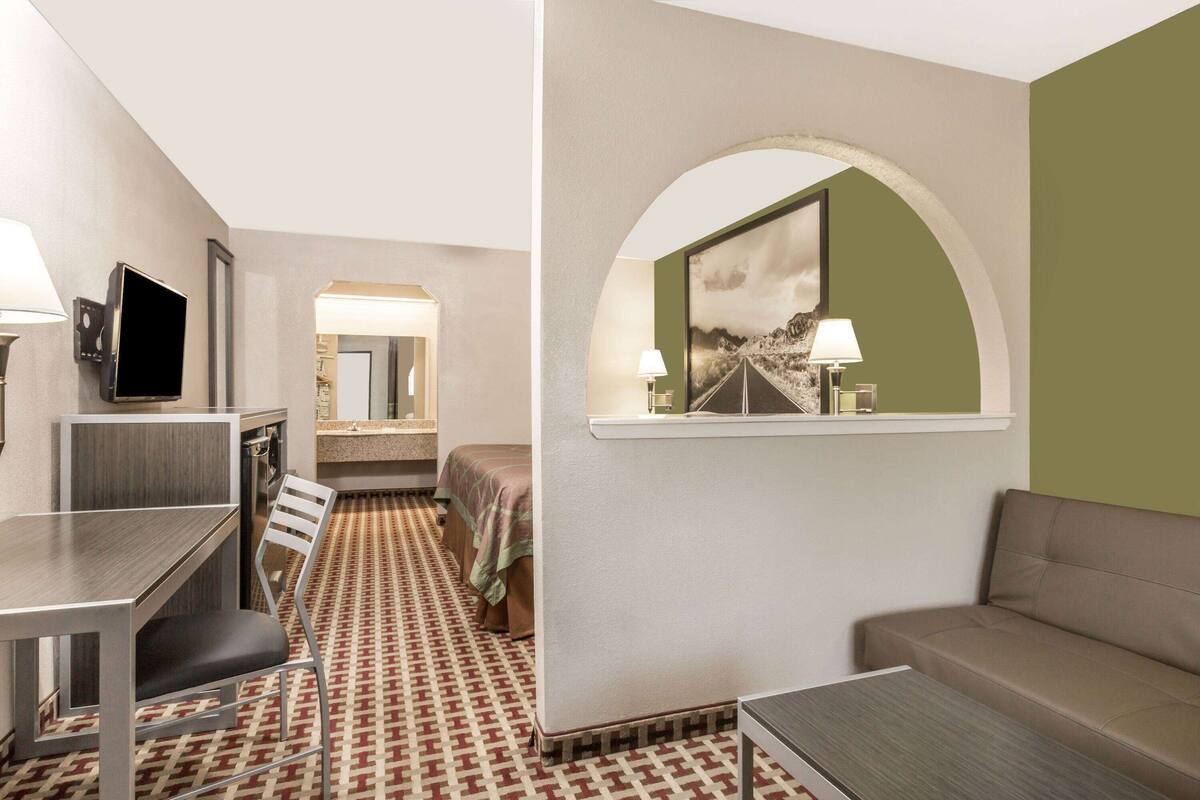 A hotel room with a bed, a desk with a chair, a sofa, a TV, and a bathroom with a mirror.