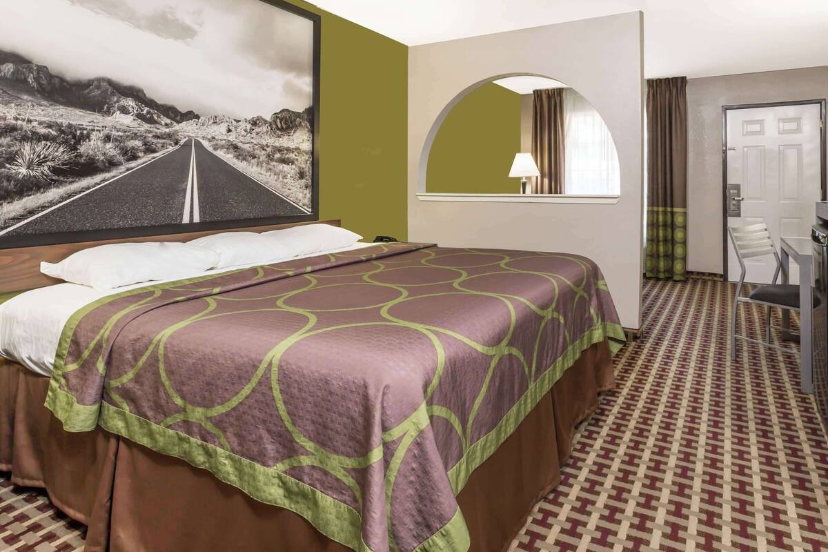 A hotel room with a bed, a large framed picture of a road, a mirror, and a desk with a chair.