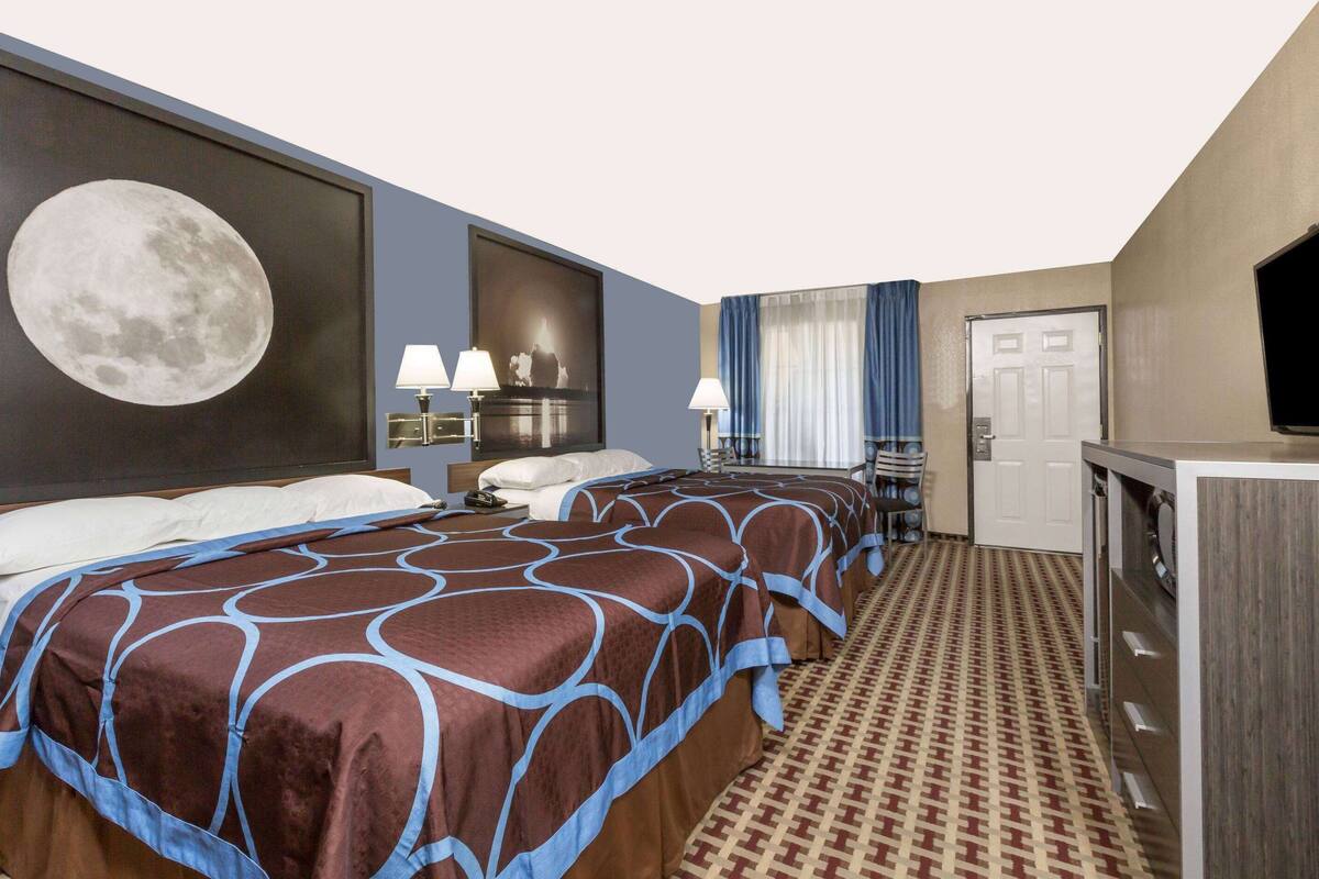 A hotel room with two beds, a large moon painting on the wall, a television, and a patterned carpet.