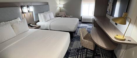 A hotel room with two beds, a desk with a lamp, a chair, a patterned rug, and a large window.