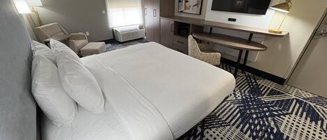 A hotel room with a large bed, a desk, a chair, a TV, and a window with blinds.