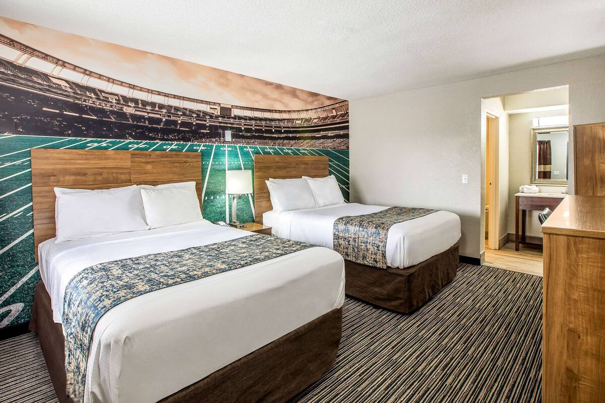 Une chambre d’hôtel avec deux lits, une grande fresque représentant un stade de football, une tête de lit en bois, une moquette à rayures et un petit bureau avec un miroir.