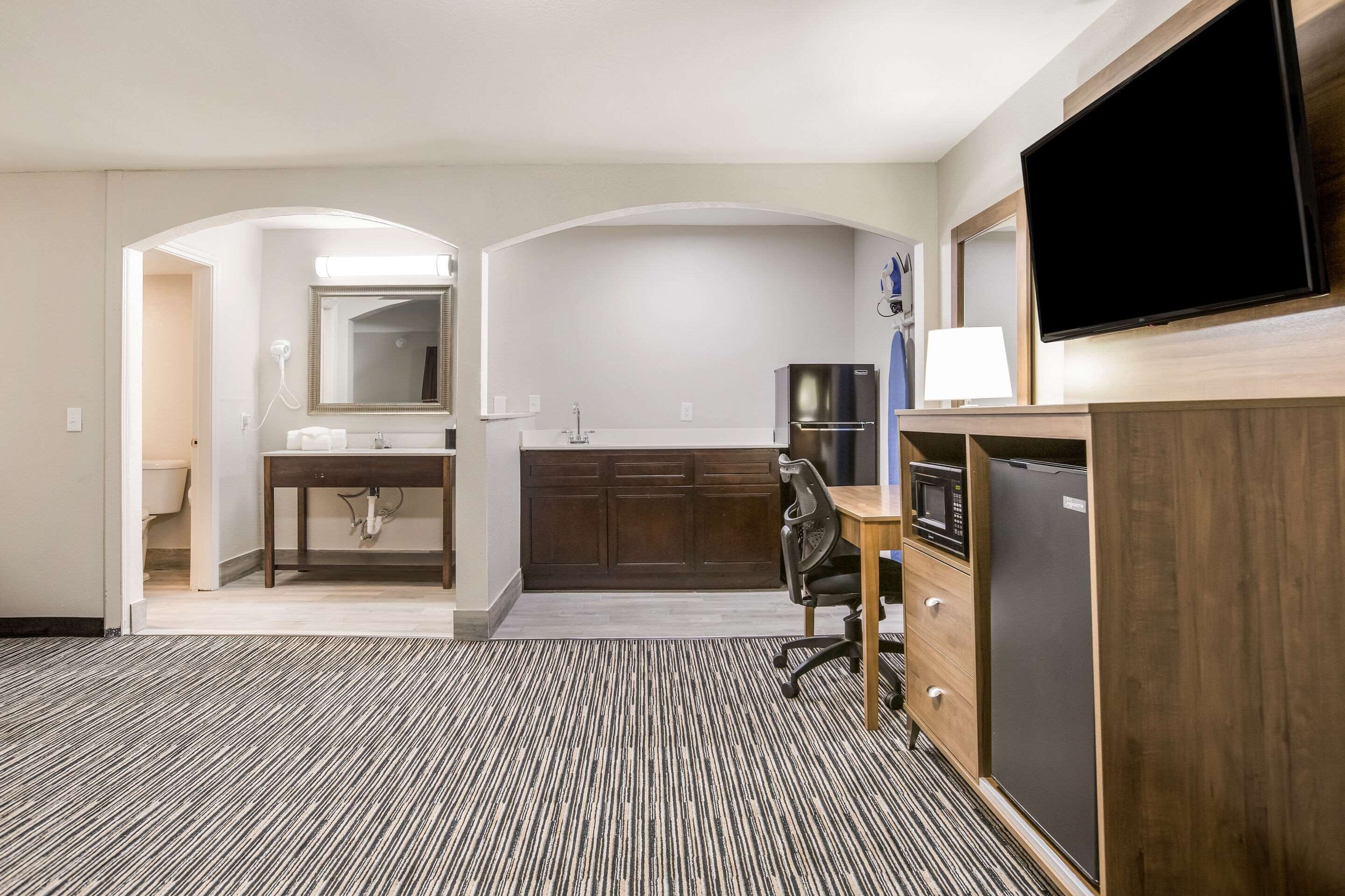 Habitación de hotel con kitchenette, televisor de pantalla plana, escritorio con silla y alfombra estampada.