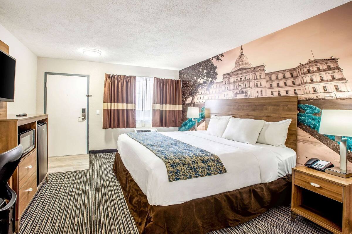 A hotel room with a bed, a desk with a computer, a TV, a window with curtains, and a wall mural.