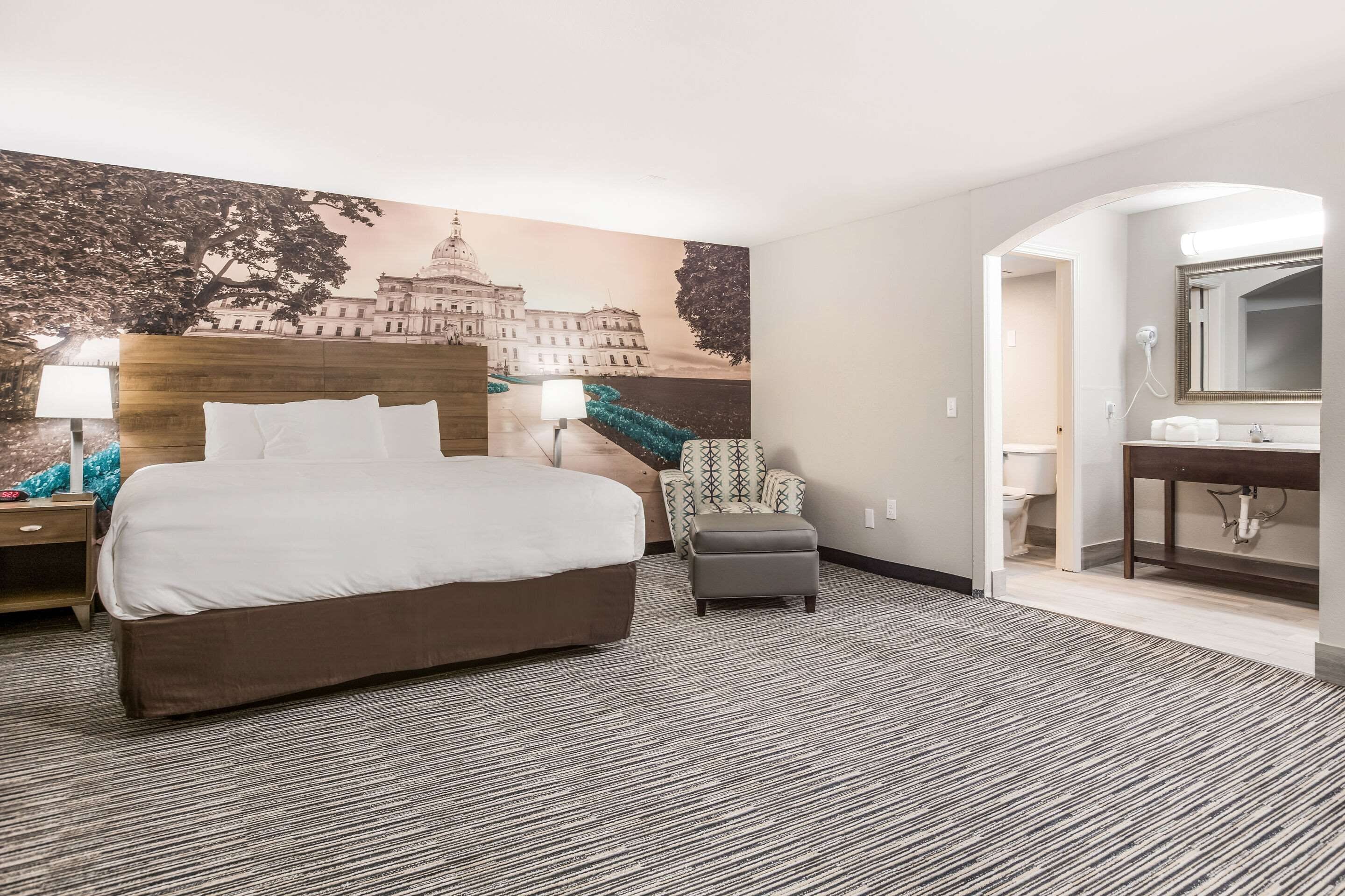 A hotel room with a large bed, a chair, a desk, and a bathroom visible through an archway.