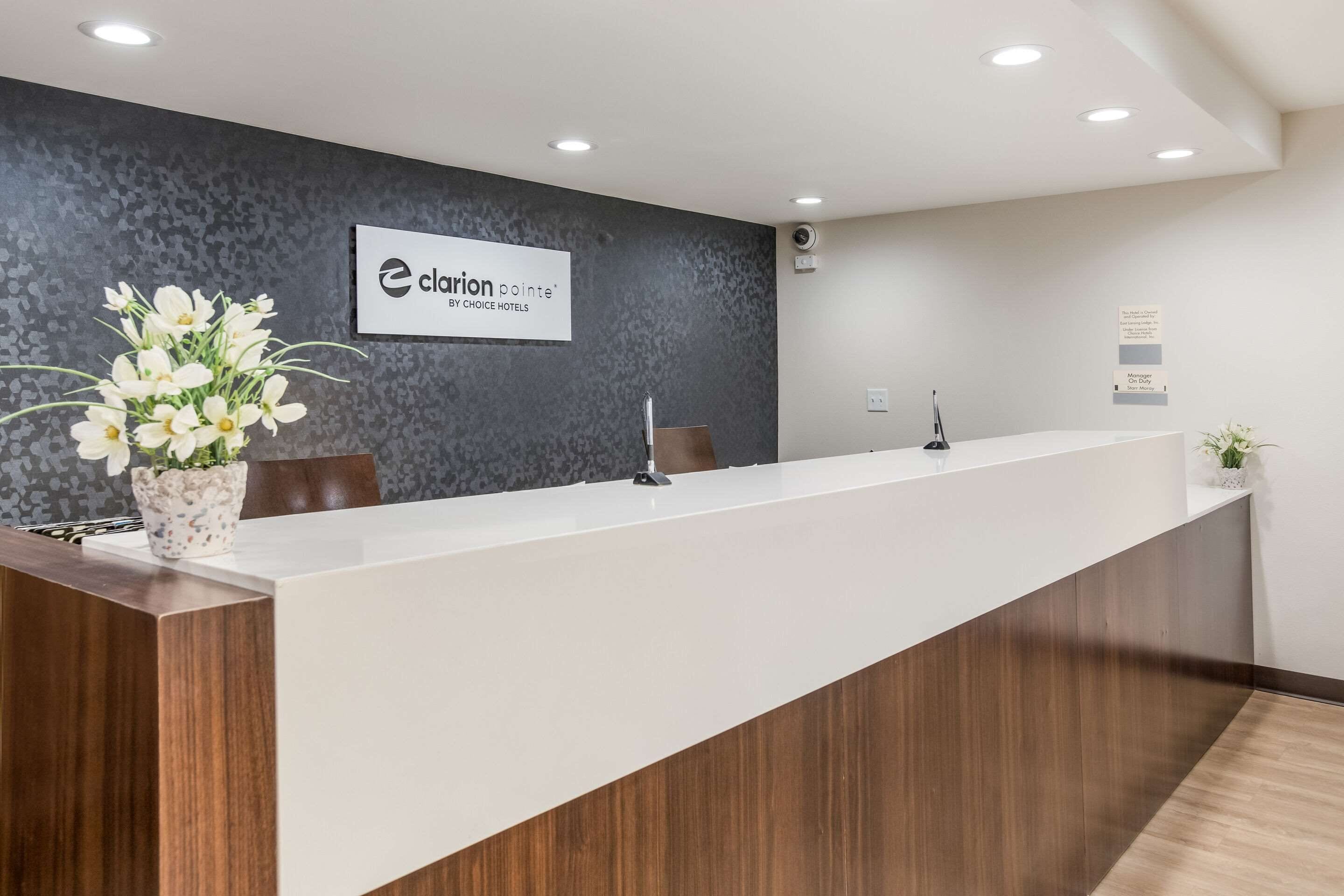 A reception desk with a dark patterned wall, a sign reading "Clarion Pointe by Choice Hotels," and a potted plant on the left.