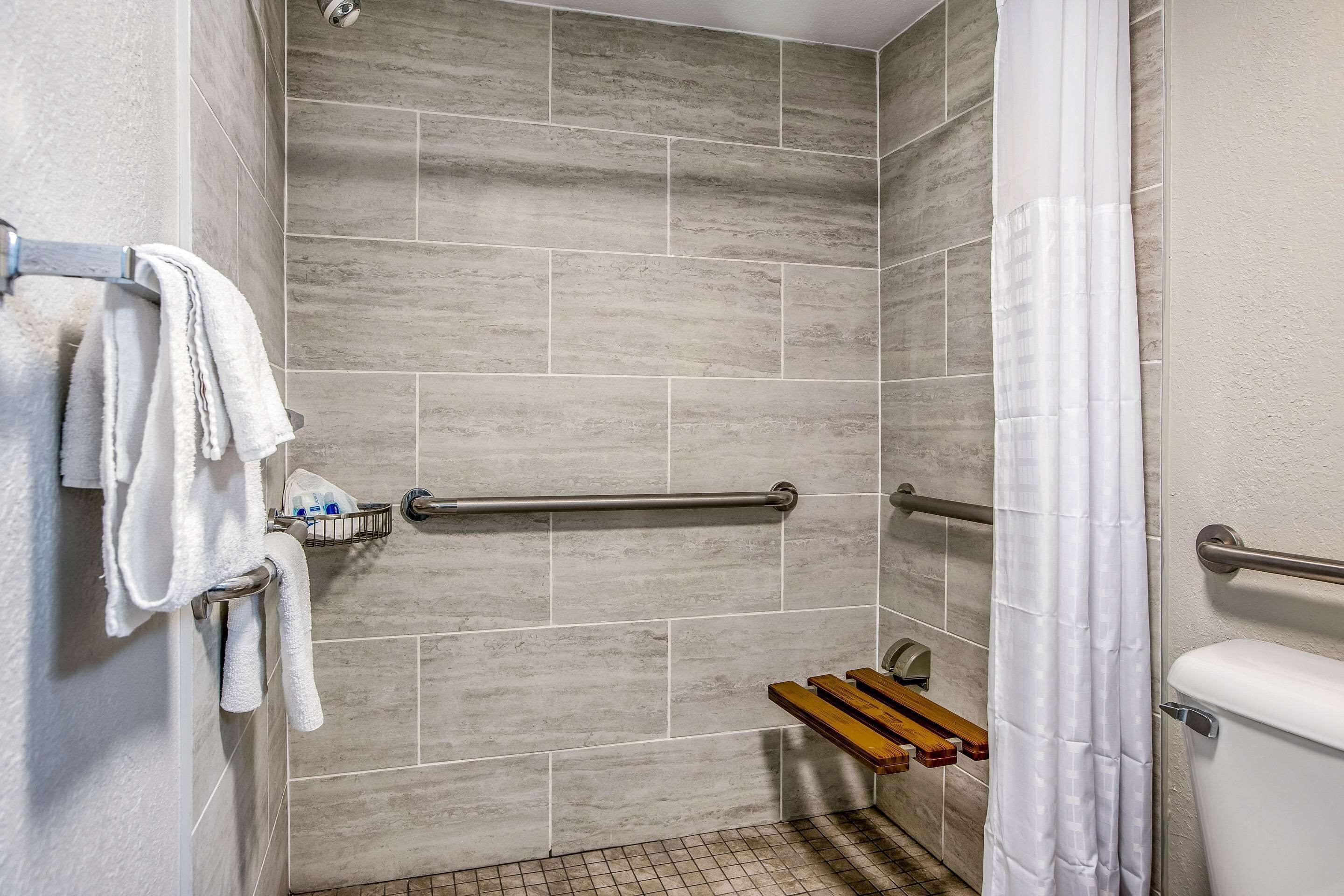A bathroom with grey tiled walls, a white toilet, a shower with a white curtain, and a grab bar.