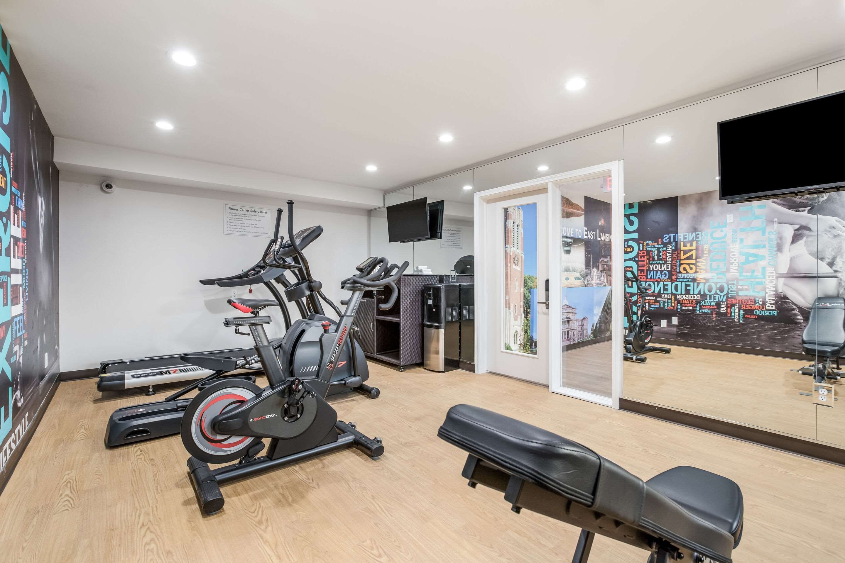 A gym with cardio machines, a bench, and a mirrored wall reflecting exercise equipment.
