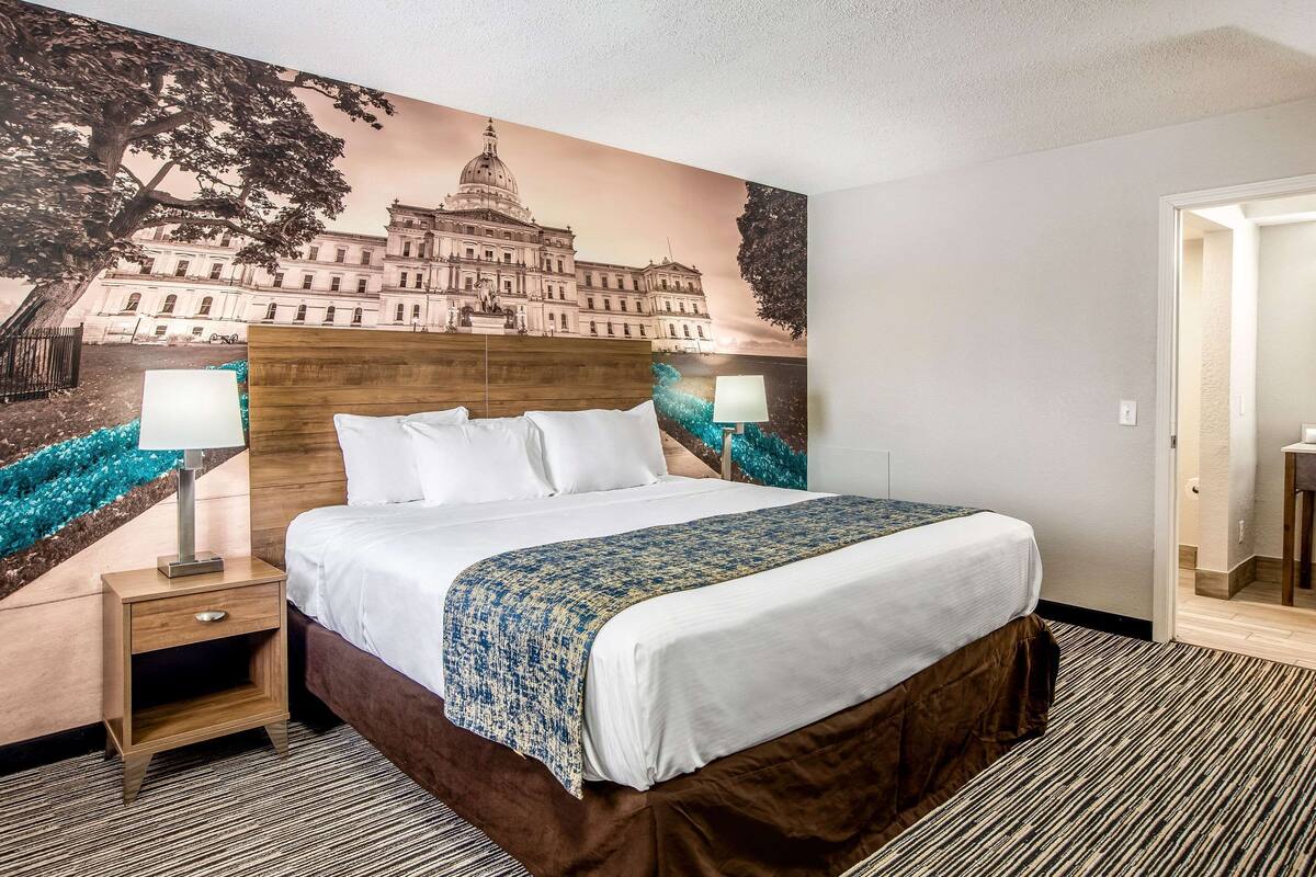 Une chambre d’hôtel avec un grand lit, deux tables de chevet avec des lampes, une fresque murale et une porte donnant sur une autre pièce.