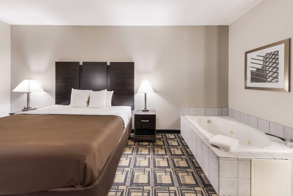 A hotel room with a bed, two bedside lamps, a nightstand, a patterned rug, a bathtub, and a framed picture on the wall.