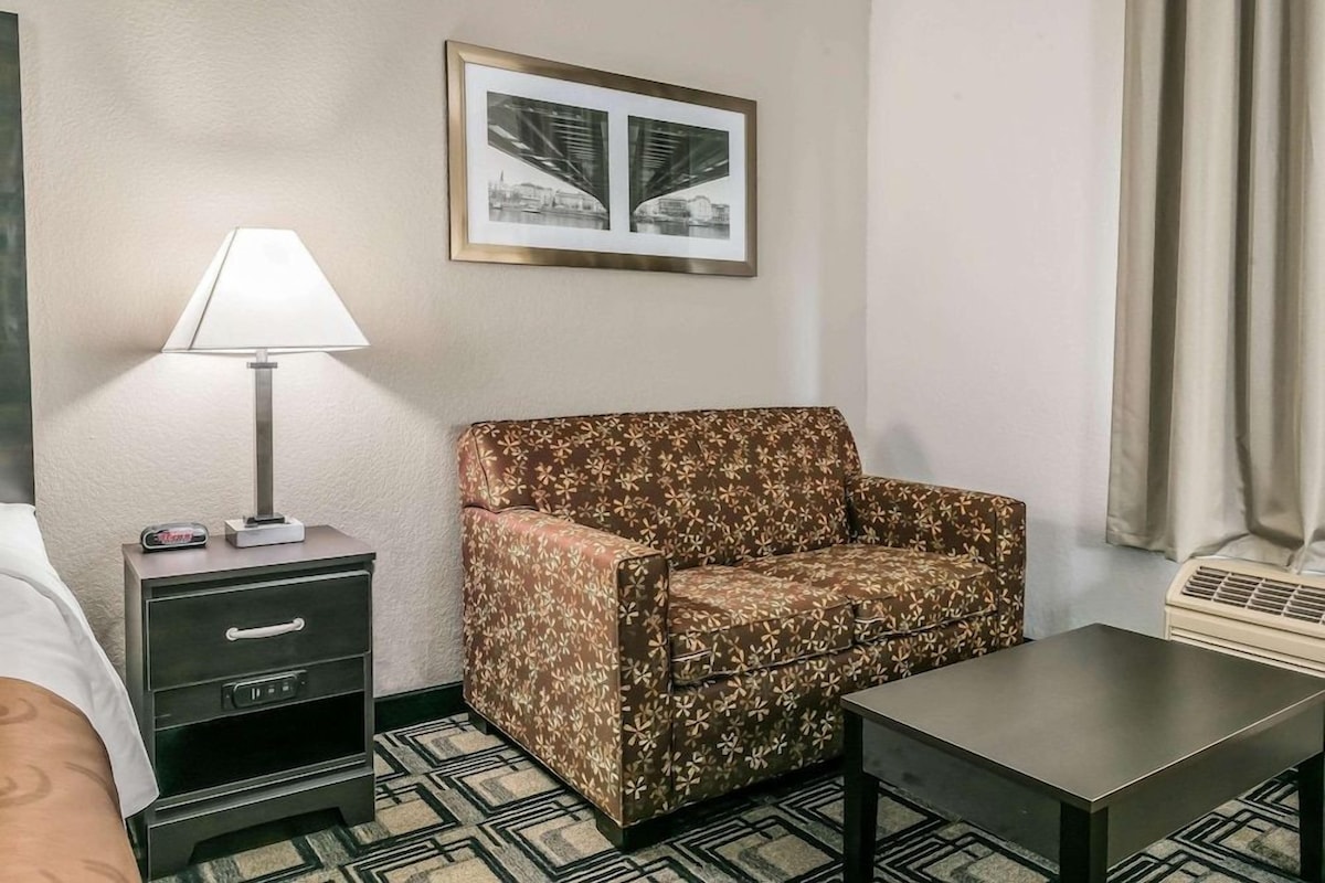 Une chambre d’hôtel avec un canapé, une table de chevet avec une lampe, un tableau encadré au mur et une fenêtre avec des rideaux.