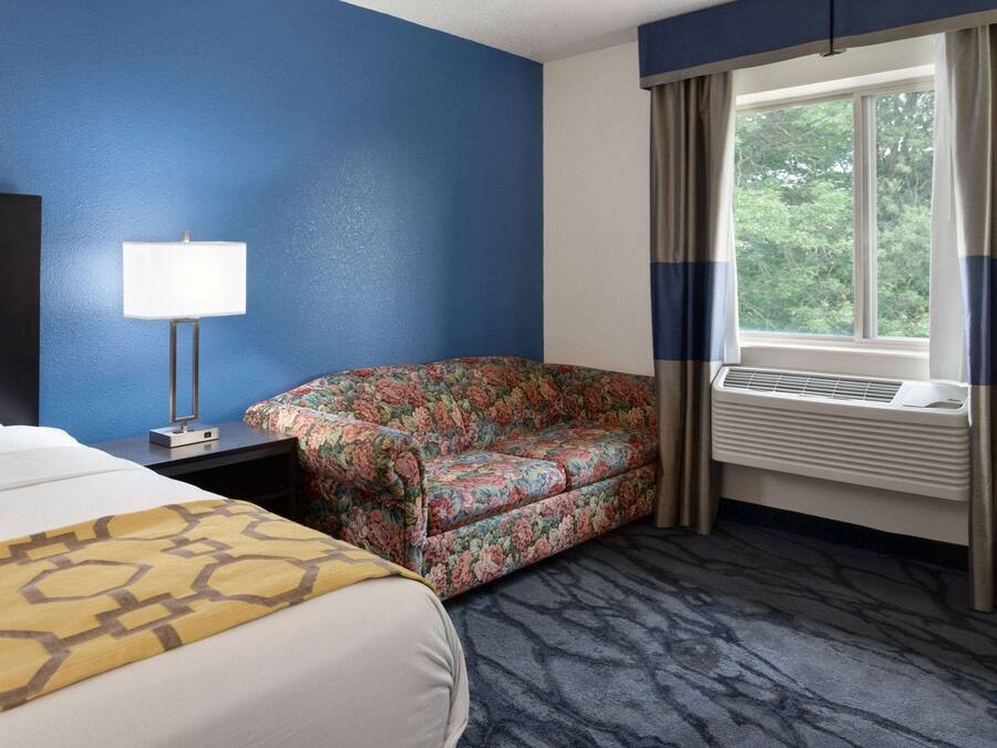 A hotel room with a bed, a floral-patterned armchair, a desk with a lamp, and a window with curtains.
