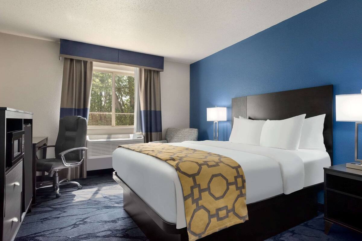 Una habitación de hotel con cama, escritorio, silla, ventana con cortinas y mesita de noche.