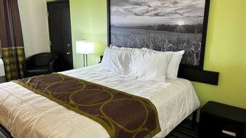 Habitación de hotel con cama, una silla, un escritorio, una lámpara y un cuadro grande con paisaje en la pared.