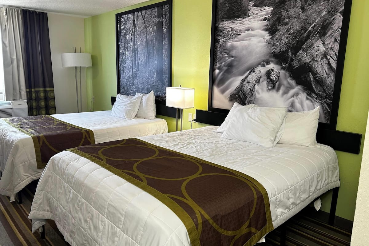 Habitación de hotel con dos camas, un amplio ventanal con cortinas, una lámpara de pared y un cuadro grande con una cascada.