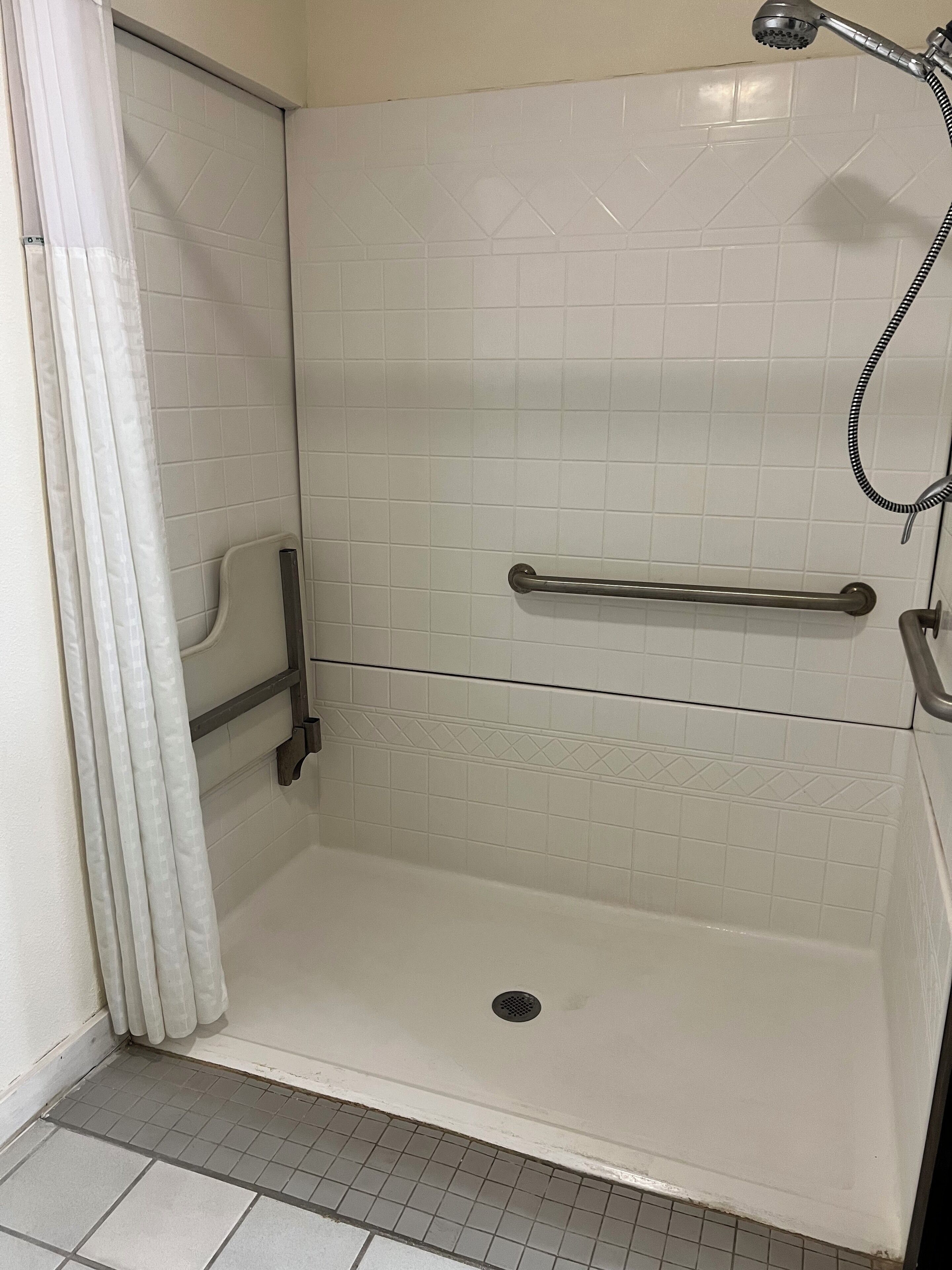 Zona de ducha con paredes de azulejos blancos, ducha de mano, barra de apoyo y desagüe en el suelo.