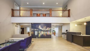 Un vestíbulo de hotel con mostrador de recepción, zona de estar con sillas moradas, una escalera de madera y un mural con vista a la ciudad.
