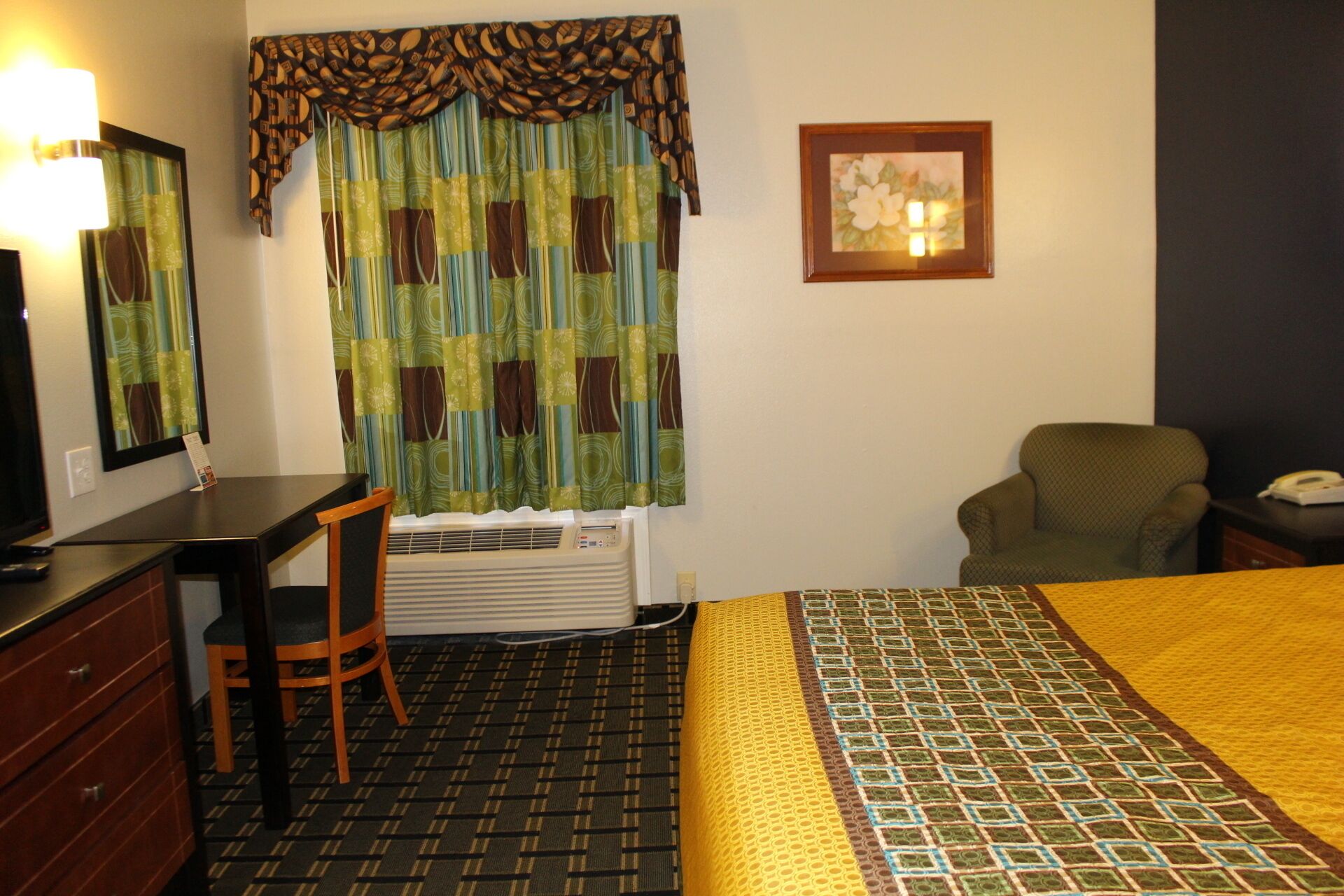 A hotel room with a bed, a desk, a chair, a heater, a lamp, and a framed picture on the wall.